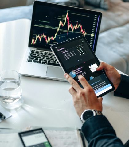 Person analyzing digital market trends on tablet with laptop showing financial charts.
